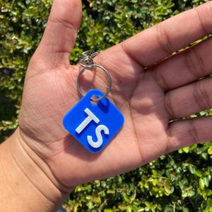 K&ouml;nnte beinhalten: Ein blauer quadratischer Schl&uuml;sselanh&auml;nger mit den wei&szlig;en Buchstaben "TS" ist an einem silbernen Schl&uuml;sselring befestigt. Der Schl&uuml;sselanh&auml;nger wird in einer Hand vor gr&uuml;nem Laub gehalten.