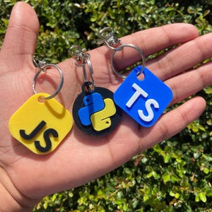 May include: Three keychains with silver rings and clasps. One yellow square keychain with "JS" in black, a black circular keychain with a blue and yellow Python logo, and a blue square keychain with "TS" in white.