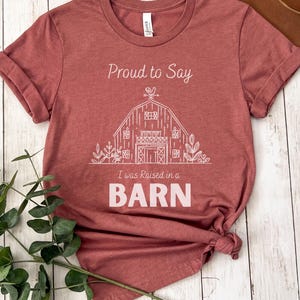May include: A maroon t-shirt with the text "Proud to Say I was Raised in a BARN" in white. The graphic is a white line drawing of a barn with a rooster on top. The shirt is folded at the bottom.