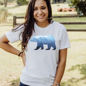 May include: A white t-shirt with a blue mountain range silhouette of a bear. The bear is outlined in black and the mountains are shaded in different shades of blue.