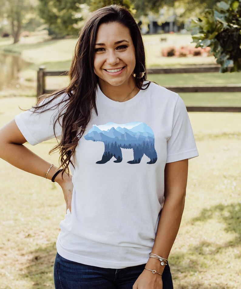 May include: A white t-shirt with a blue mountain range silhouette of a bear. The bear is outlined in black and the mountains are shaded in different shades of blue.