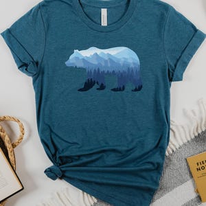 May include: A teal blue t-shirt with a silhouette of a bear. The bear is filled with a blue and white mountain landscape.