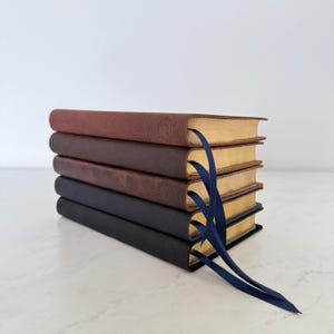 May include: A stack of five leather-bound books in varying shades of brown and black. Each book has a gold-colored page edge and a blue ribbon bookmark. The books are arranged on a white surface.