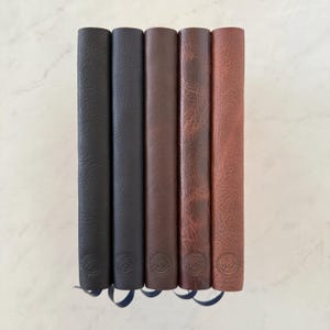 Può includere: Cinque diari verticali con copertine testurizzate disposti fianco a fianco. I colori sfumano dal nero al marrone scuro, marrone medio, marrone rossiccio e beige chiaro. Ogni diario presenta un piccolo logo circolare in rilievo sul fondo.