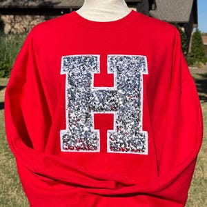 May include: A red sweatshirt with a large, sequined letter "H" in silver. The letter is centered on the front of the long-sleeved crew neck top. The background shows a house and some greenery.