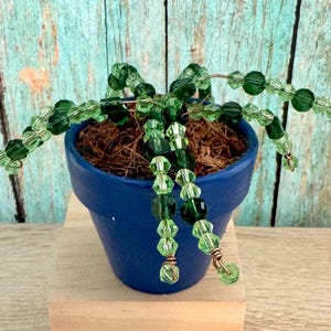 May include: A small, blue painted ceramic pot filled with brown material, holding a beaded plant. The plant features green beads strung on wire, creating a decorative, handcrafted piece. The background is a weathered, turquoise-colored wooden surface.