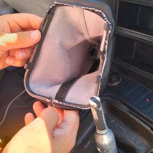 Puede incluir: Fuelle de palanca de cambios de cuero negro con interior gris, abierto. La palanca de cambios es visible, junto con partes del interior del coche, incluyendo el salpicadero y la consola central. El fuelle es probablemente para una transmisión manual.