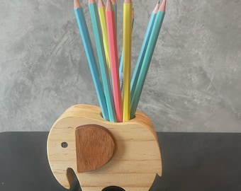 May include: A wooden elephant-shaped pencil holder with a brown ear detail, holding several pencils in various colors. The pencils are blue, yellow, and pink. The holder is made of light-colored wood.