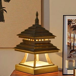 May include: A wooden pagoda-style table lamp with a tiered roof design. The lamp has a square base and a warm, inviting glow emanating from within. The lamp sits on a dark wooden surface, with a framed picture in the background.