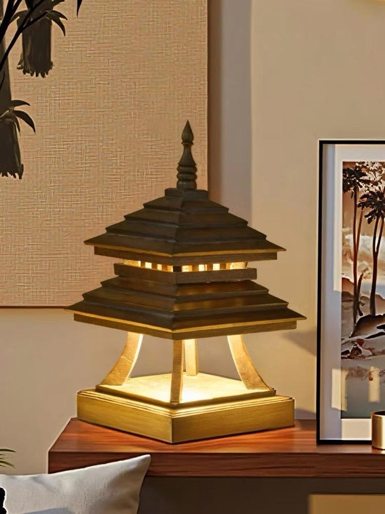 May include: A wooden pagoda-style table lamp with a tiered roof design. The lamp has a square base and a warm, inviting glow emanating from within. The lamp sits on a dark wooden surface, with a framed picture in the background.