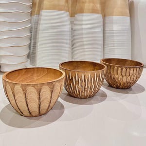 Puede incluir: Tres cuencos de madera con diseños tallados. Los cuencos son de madera de color marrón claro y tienen diferentes patrones, incluyendo ranuras verticales, óvalos alargados y una textura similar a un panal. Los cuencos se exhiben sobre una superficie blanca.