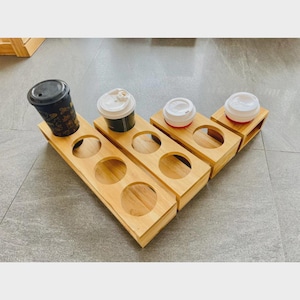 Może przedstawiać: Drewniane uchwyty na kubki do kawy z okrągłymi wycięciami, każdy z innym kubkiem do kawy. Kubki są czarne i białe z pokrywkami. Uchwyty wykonane są z jasnego drewna i ułożone w rzędzie.