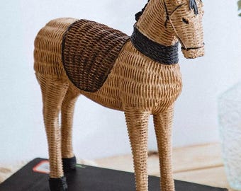 Puede incluir: Una figurita de caballo de mimbre tejido con una silla de montar marrón y detalles negros. El caballo está de pie sobre un libro.