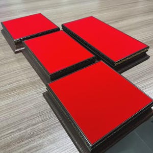 Puede incluir: Cuatro posavasos rectangulares con superficies rojas brillantes y bordes marrones oscuros. Los posavasos están dispuestos sobre una superficie marrón claro, mostrando un diseño moderno. Los bordes tienen un ribete dorado.