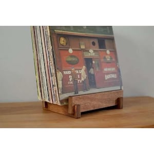 May include: A wooden record holder displaying a collection of vinyl records. The holder is made of light brown wood and has a simple, rectangular design. The record covers feature a vintage bar scene with the words "Cocktails" and "Hard Rock Cafe".