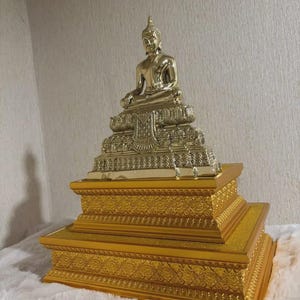 Peut inclure: Une statue dorée de Bouddha assise sur une base ornée à plusieurs niveaux. La statue est ornée de sculptures complexes et d'une expression sereine. La base est de couleur or riche, ajoutant à l'apparence royale de la statue.