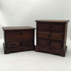 Op de afbeelding: Twee houten opbergkisten. Eén is een kleine kist met een scharnierend deksel en twee kleine lades. De andere is een kleine dressoir met drie lades. Beide zijn gemaakt van donker hout en hebben ronde knoppen.