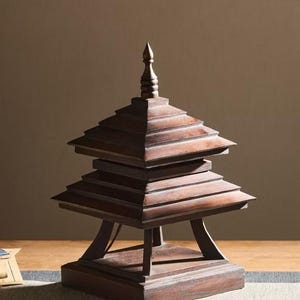 May include: A wooden pagoda-style sculpture with a tiered roof and a pointed top. The structure is made of dark brown wood and has a square base. The sculpture is displayed on a wooden surface.