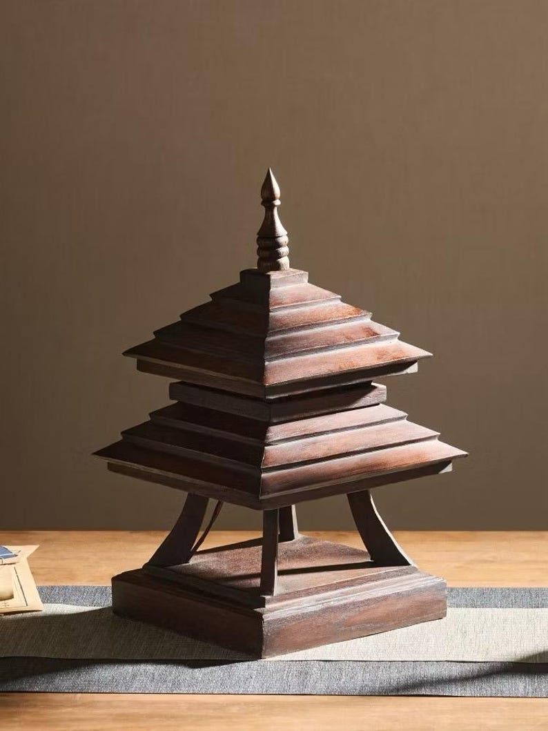 May include: A wooden pagoda-style sculpture with a tiered roof and a pointed top. The structure is made of dark brown wood and has a square base. The sculpture is displayed on a wooden surface.