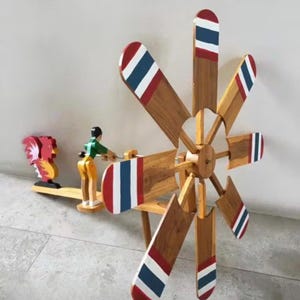 Puede incluir: Una escultura cinética de madera con una gran rueda de paletas con detalles en rojo, blanco y azul en las aspas. Una pequeña figura está colocada cerca de la rueda, y una figura de gallo está en un balancín. La escultura es de madera.