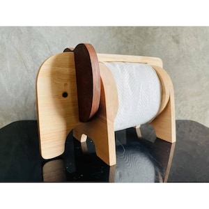 May include: A wooden elephant-shaped paper towel holder. The holder is made of light-coloured wood with a darker brown accent for the ear. A roll of white paper towels is visible inside the holder. The design is whimsical and functional.