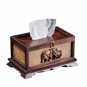 May include: A rectangular wooden tissue box with a dark brown finish. The front features a woven pattern and a carved relief of three elephants. A stack of white tissues protrudes from the top.