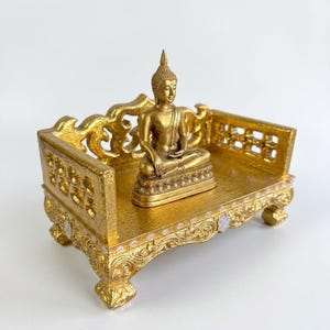 Peut inclure: Une figurine de Bouddha dorée assise en position de méditation sur une plateforme rectangulaire ornée. La plateforme présente des détails sculptés complexes, des panneaux latéraux ajourés et de petites fleurs blanches sur le bord.