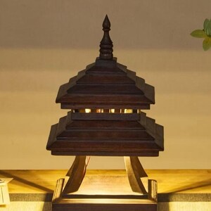 May include: A dark brown wooden pagoda-style lamp with a tiered roof and a pointed finial. The lamp has a rectangular base and is supported by angled wooden supports. The lamp is lit from within, casting a warm glow.