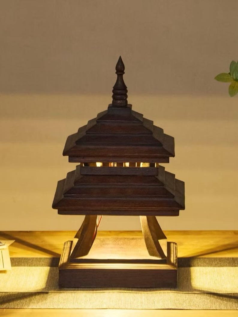 May include: A dark brown wooden pagoda-style lamp with a tiered roof and a pointed finial. The lamp has a rectangular base and is supported by angled wooden supports. The lamp is lit from within, casting a warm glow.