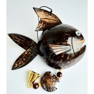 May include: A handcrafted, dark brown fish-shaped wind chime made from coconut shell. The fish has a round body, fins, and a small bell and bird-shaped charm hanging below. The fish has a white eye and a black string for hanging.