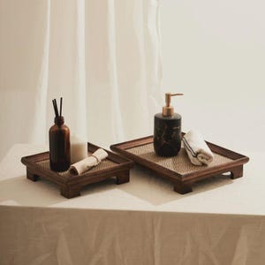 Puede incluir: Dos bandejas de madera con inserciones tejidas. Una bandeja contiene una botella de vidrio marrón con cañas, una vela blanca y una toalla blanca enrollada. La otra tiene un dispensador de jabón de mármol negro y una toalla blanca doblada.