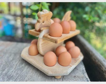 May include: A wooden egg holder shaped like a chicken, holding a dozen brown eggs. The two-tiered design features a chicken head and wing details. The holder is made of light-colored wood and sits on a wooden surface.