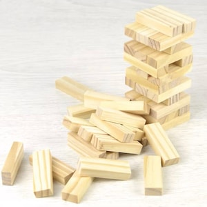 Puede incluir: Un juego de apilamiento de bloques de madera con una torre de bloques parcialmente construida. Varios bloques están esparcidos alrededor de la torre. Los bloques son de color marrón claro y rectangulares.