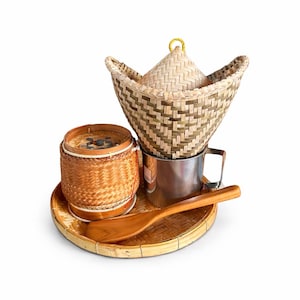 Może przedstawiać: Zestaw tradycyjnych azjatyckich narzędzi kuchennych. Zestaw zawiera tkany parownik bambusowy, mały okrągły kosz, drewnianą łyżkę i garnek ze stali nierdzewnej, wszystko ułożone na tkanym tacy. Przedmioty wykonane są z naturalnych materiałów.