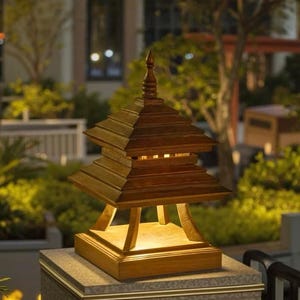 May include: A wooden pagoda-style outdoor lamp with a tiered roof and a pointed finial. The lamp is illuminated from within, casting a warm glow. It sits atop a stone pillar with a gold trim, set against a blurred background of greenery.