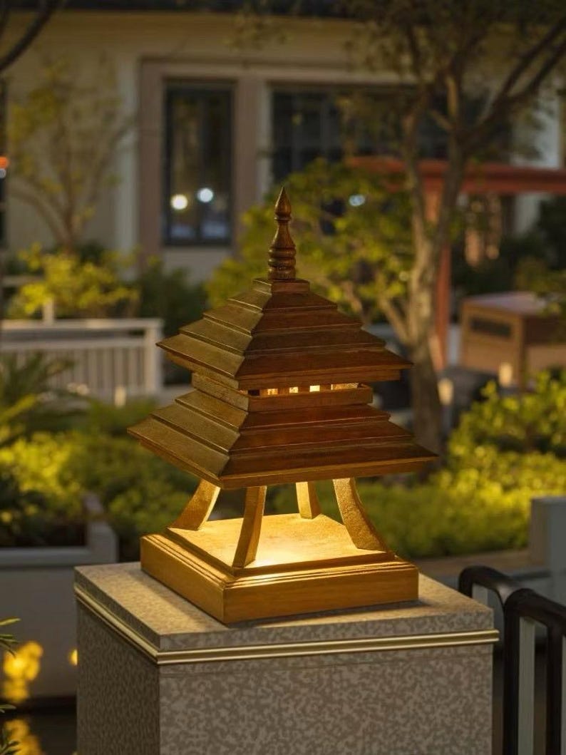 May include: A wooden pagoda-style outdoor lamp with a tiered roof and a pointed finial. The lamp is illuminated from within, casting a warm glow. It sits atop a stone pillar with a gold trim, set against a blurred background of greenery.