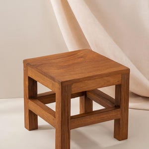 May include: A small, wooden stool with a square top and a simple, open frame design. The stool is made of light brown wood and has a natural finish.