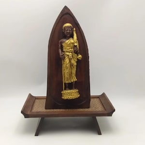 K&ouml;nnte beinhalten: Holzschnitzerei einer goldfarbenen Figur in Robe, die einen Stab und eine Schale h&auml;lt. Die Figur ist vor einem dunkelbraunen, gew&ouml;lbten Holz-Hintergrund platziert. Die Schnitzerei wird auf einem kleinen, niedrigen Holztisch mit gewebter Oberfl&auml;che ausgestellt.