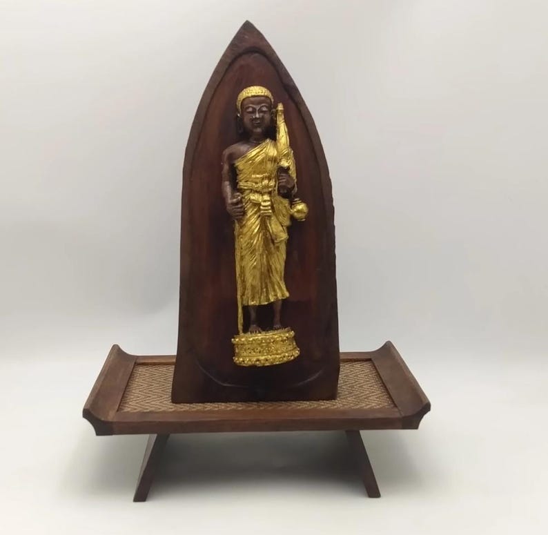 K&ouml;nnte beinhalten: Holzschnitzerei einer goldfarbenen Figur in Robe, die einen Stab und eine Schale h&auml;lt. Die Figur ist vor einem dunkelbraunen, gew&ouml;lbten Holz-Hintergrund platziert. Die Schnitzerei wird auf einem kleinen, niedrigen Holztisch mit gewebter Oberfl&auml;che ausgestellt.