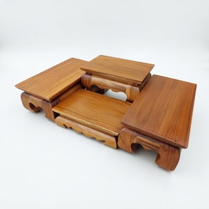 Puede incluir: Un expositor de madera de varios niveles con cuatro plataformas rectangulares de diferentes alturas. La veta de la madera marrón natural es visible en todas las superficies, sostenido por patas talladas con recortes curvos.