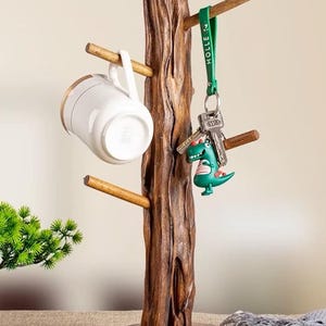 Può includere: Un porta tazze e chiavi in legno a forma di albero con una tazza bianca, un portachiavi di dinosauro verde e un portachiavi verde con la scritta "HOLLE" su di esso.
