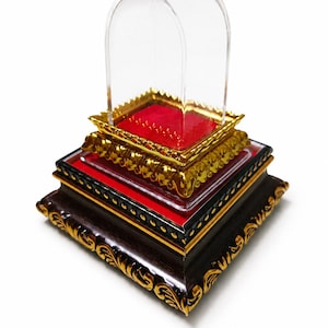 May include: A tiered display stand featuring a dark brown square base with gold filigree accents, a red velvet platform, and a smaller gold-toned square frame with a clear arched acrylic cover. The structure is designed for showcasing small items.