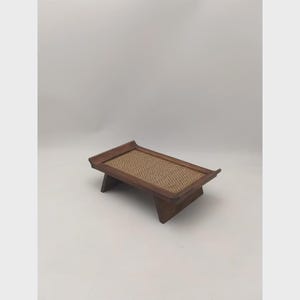 K&ouml;nnte beinhalten: Ein kleiner, rechteckiger Holztablett mit einer gewebten, hellbraunen Oberfl&auml;che. Das Tablett hat einen dunkelbraunen Rahmen und abgewinkelte Beine. Das Design ist schlicht und minimalistisch, geeignet zum Servieren oder Ausstellen.
