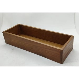 Puede incluir: Una caja de madera rectangular con un acabado marrón natural. La caja está vacía y tiene una superficie lisa.