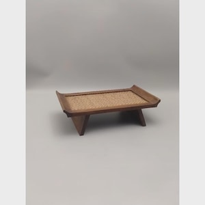 K&ouml;nnte beinhalten: Ein Serviertablett aus Holz mit einem Einsatz aus geflochtenem Rattan. Das Tablett hat eine rechteckige Form mit leicht gebogenen Kanten und wird von vier abgewinkelten Beinen getragen. Das Holz hat einen warmen, nat&uuml;rlichen Farbton.