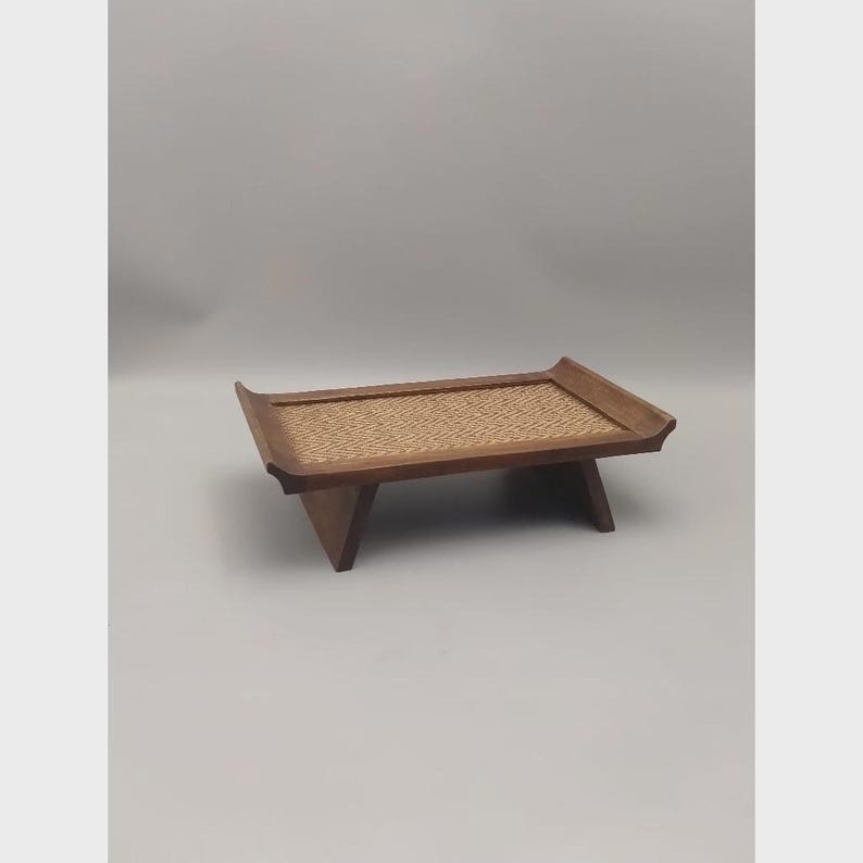 K&ouml;nnte beinhalten: Ein Serviertablett aus Holz mit einem Einsatz aus geflochtenem Rattan. Das Tablett hat eine rechteckige Form mit leicht gebogenen Kanten und wird von vier abgewinkelten Beinen getragen. Das Holz hat einen warmen, nat&uuml;rlichen Farbton.