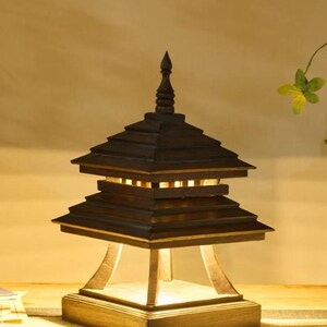 May include: A wooden pagoda-style lamp with a dark brown finish. The lamp has a tiered roof design and a square base. The light shines through the openings in the roof. The lamp is a decorative lighting fixture.