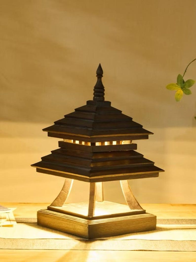 May include: A wooden pagoda-style lamp with a dark brown finish. The lamp has a tiered roof design and a square base. The light shines through the openings in the roof. The lamp is a decorative lighting fixture.