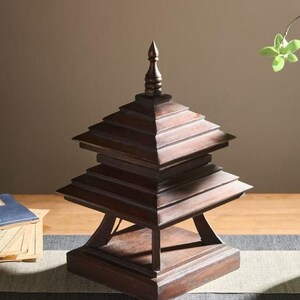 May include: A dark brown, tiered wooden pagoda-style decorative object. The pagoda has a square base and multiple layers, each slightly smaller than the one below. The top has a pointed finial. The object is on a wooden table.