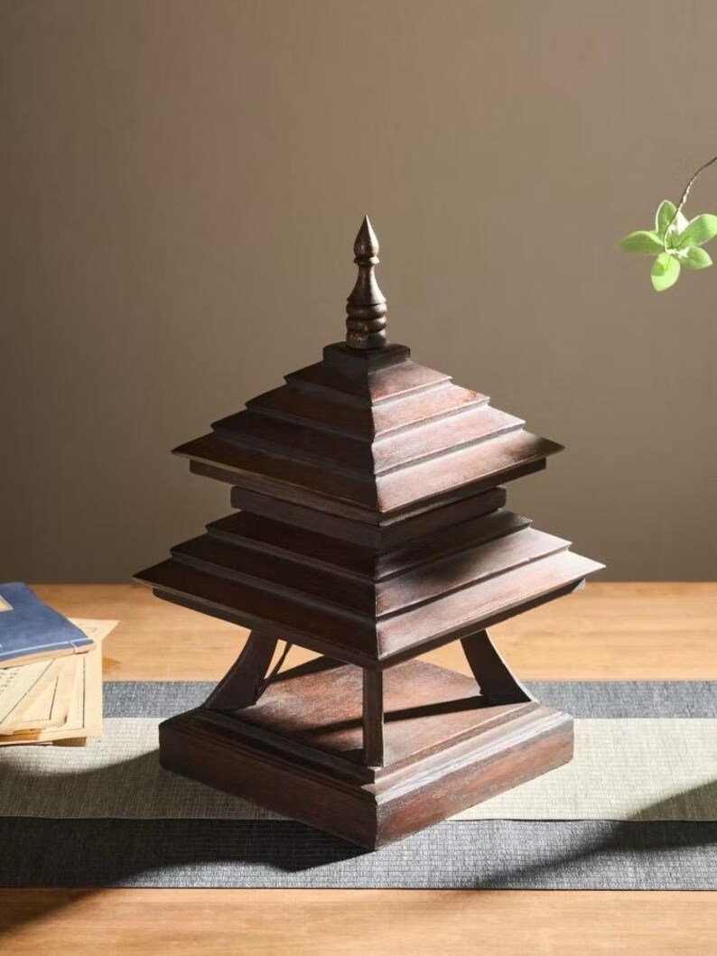 May include: A dark brown, tiered wooden pagoda-style decorative object. The pagoda has a square base and multiple layers, each slightly smaller than the one below. The top has a pointed finial. The object is on a wooden table.